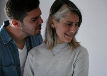 Homem sussurrando agressivamente no ouvido de uma mulher que chora e tenta se afastar, expressando angústia e sofrimento emocional.