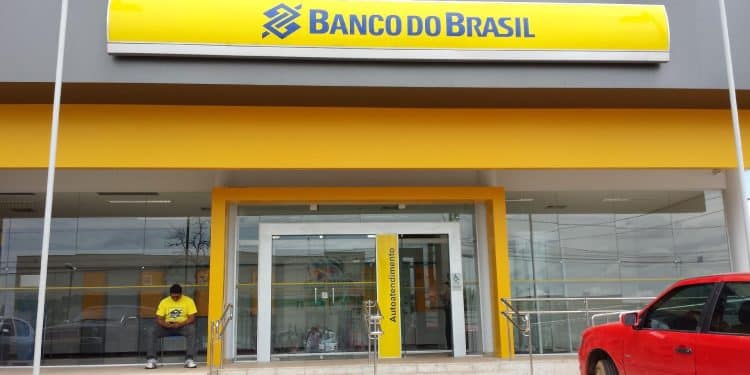 Homem de camisa amarela sentado na entrada de agência do Banco do Brasil moderna