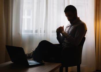 Homem sentado sozinho em cadeira com laptop aberto em ambiente escuro representando os riscos de viver isolado para a saúde física e mental