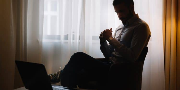 Homem sentado sozinho em cadeira com laptop aberto em ambiente escuro representando os riscos de viver isolado para a saúde física e mental