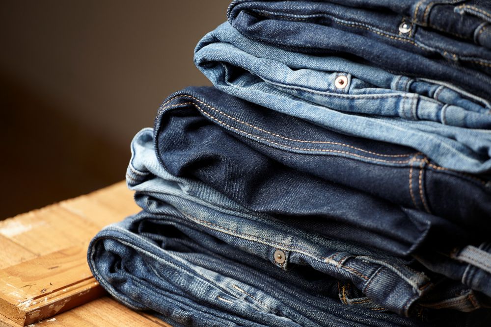 Várias calças jeans dobradas e empilhadas sobre uma mesa de madeira rústica, exibindo diferentes lavagens e texturas de denim.