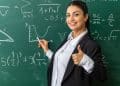 Professora sorridente vestindo blazer preto e camisa branca, apontando para um gráfico em um quadro negro repleto de equações matemáticas e fórmulas.