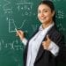Professora sorridente vestindo blazer preto e camisa branca, apontando para um gráfico em um quadro negro repleto de equações matemáticas e fórmulas.