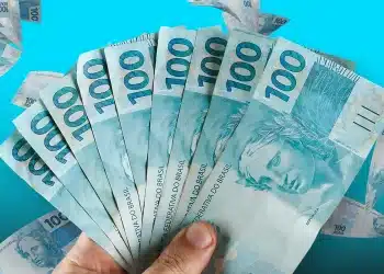 Mão segurando leque de notas de cem reais em fundo azul vibrante