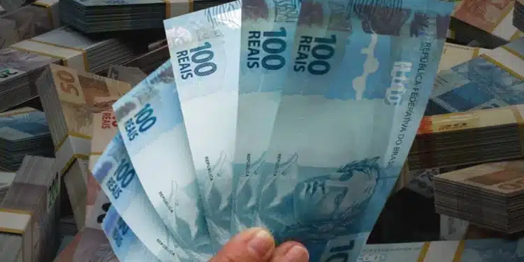 Mão segurando várias notas de R$ 100 com maços de dinheiro ao fundo, representando o valor disponível para saque no saque-aniversário do FGTS 2026, cujo prazo para nascidos em janeiro encerra em 31 de março de 2026