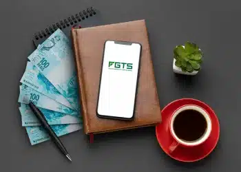 Celular com o logo do FGTS apoiado em uma agenda, ao lado de notas de R$ 100, caneta e xícara de café, simbolizando o planejamento financeiro para o saque-aniversário
