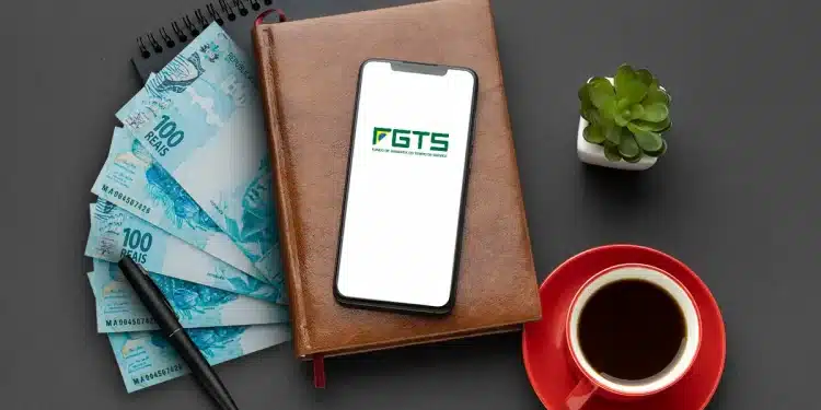 Celular com o logo do FGTS apoiado em uma agenda, ao lado de notas de R$ 100, caneta e xícara de café, simbolizando o planejamento financeiro para o saque-aniversário
