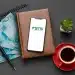 Celular com o logo do FGTS apoiado em uma agenda, ao lado de notas de R$ 100, caneta e xícara de café, simbolizando o planejamento financeiro para o saque-aniversário