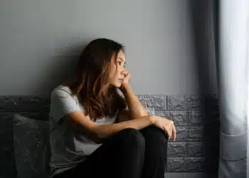 Mulher sentada sozinha olhando pela janela com expressão pensativa representando sinais de sofrimento emocional e alerta para a saúde mental.