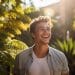 Retrato de um homem jovem de cabelos cacheados, sorrindo de forma genuína e relaxada, com o rosto voltado para a luz do sol, rodeado por folhagens verdes em um jardim ensolarado.