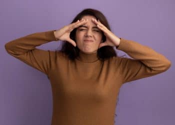 Mulher com expressão de desconforto, com as mãos na cabeça, mostrando sinais de tensão emocional, usando um suéter marrom, com fundo roxo.