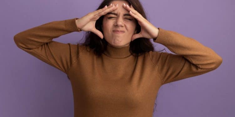 Mulher com expressão de desconforto, com as mãos na cabeça, mostrando sinais de tensão emocional, usando um suéter marrom, com fundo roxo.