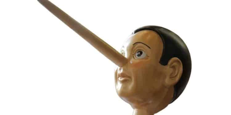 Fotografia em close de uma estatueta de madeira do personagem Pinóquio, destacando o nariz comprido e a expressão surpresa, isolada contra um fundo branco e limpo, servindo como ilustração visual para o conceito de mentira.