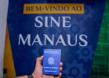 Mão segurando carteira de trabalho em frente ao banner do Sine Manaus