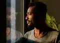 Homem olhando pela janela com expressão introspectiva representando sentimentos de solidão e isolamento emocional.