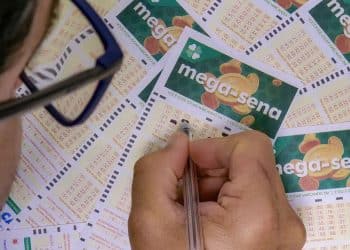 Close-up de uma pessoa preenchendo um volante da Mega-Sena com uma caneta, rodeada por outros cartões de apostas sobre uma mesa.