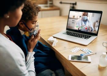 Mãe e filha durante consulta médica online, com a criança usando máscara de oxigênio enquanto recebe atendimento de um pediatra virtual.
