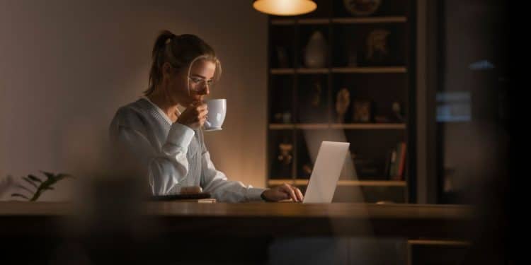 Close de uma jovem loira de óculos e colete de tricô cinza sobre camisa branca, sentada à mesa. Ela está bebendo de uma xícara branca enquanto usa um notebook, iluminada por uma luminária de mesa em um ambiente noturno aconchegante com estantes ao fundo.