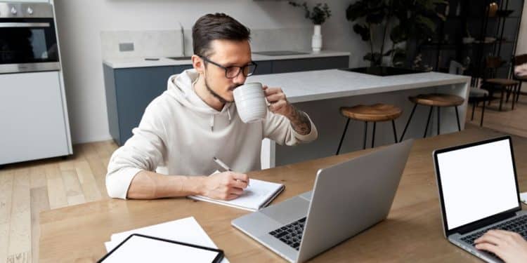 Homem jovem de óculos e moletom claro sentado à mesa de madeira, bebendo café enquanto escreve em um caderno e olha para a tela de um notebook aberto em ambiente doméstico moderno.