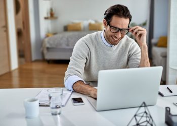 Homem sorridente de óculos trabalhando em um laptop branco em sua mesa de escritório em casa, com um quarto ao fundo.