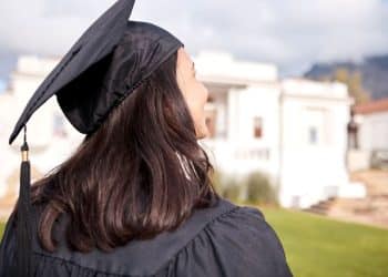 Vista traseira de uma jovem com cabelos escuros vestindo beca e capelo de formatura, olhando para um prédio clássico universitário ao fundo em um dia ensolarado.