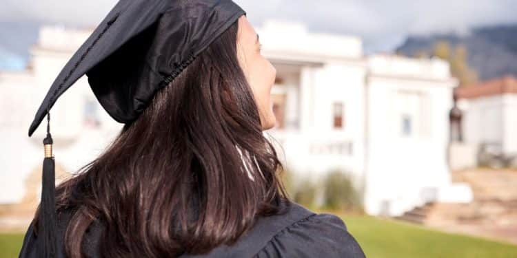 Vista traseira de uma jovem com cabelos escuros vestindo beca e capelo de formatura, olhando para um prédio clássico universitário ao fundo em um dia ensolarado.
