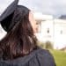 Vista traseira de uma jovem com cabelos escuros vestindo beca e capelo de formatura, olhando para um prédio clássico universitário ao fundo em um dia ensolarado.