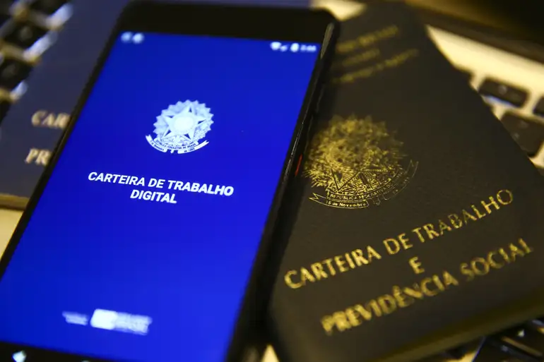 Um smartphone exibindo o aplicativo Carteira de Trabalho Digital ao lado de uma Carteira de Trabalho e Previdência Social física sobre um teclado de notebook.