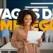 Mulher negra sorridente com cabelos cacheados, vestindo camisa bege, segurando folhas de papel em um escritório moderno com a sobreposição de texto "VAGAS DE EMPREGO".