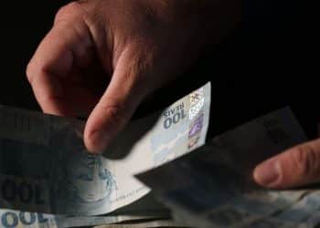 Mãos contando cédulas de R$ 100 reais — como calcular o valor do 13º salário do INSS em 2026