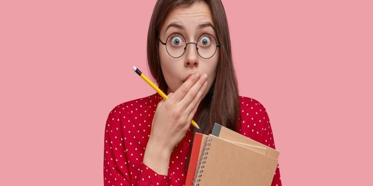 Mulher de óculos com expressão de surpresa cobrindo a boca com a mão, segurando cadernos e um lápis, representando a reação de quem descobre que pronuncia palavras erradas no português