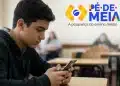Estudante do ensino médio usando celular em sala de aula, com o logo do programa Pé-de-Meia ao lado, representando o aviso do MEC para alunos que não acessaram o benefício.