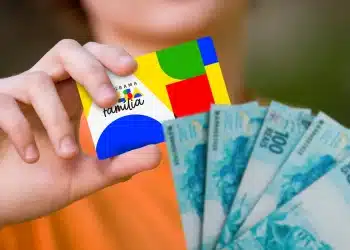 Cartão do Bolsa Família 2026 em mãos, com notas de R$ 100 ao fundo.