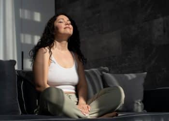 Mulher de cabelos cacheados escuros, regata branca e calça verde, sentada em sofá cinza com olhos fechados.