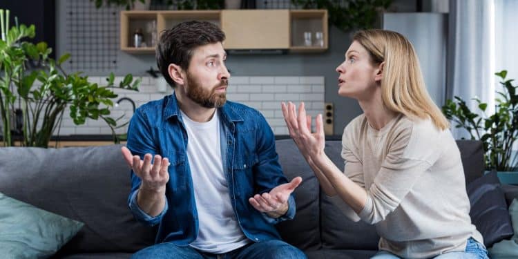 Casal discute gesticulando em sofá cinza na sala de estar.