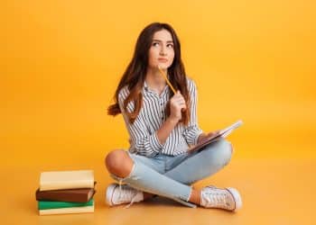 Jovem pensativa com caderno e lápis em fundo laranja.