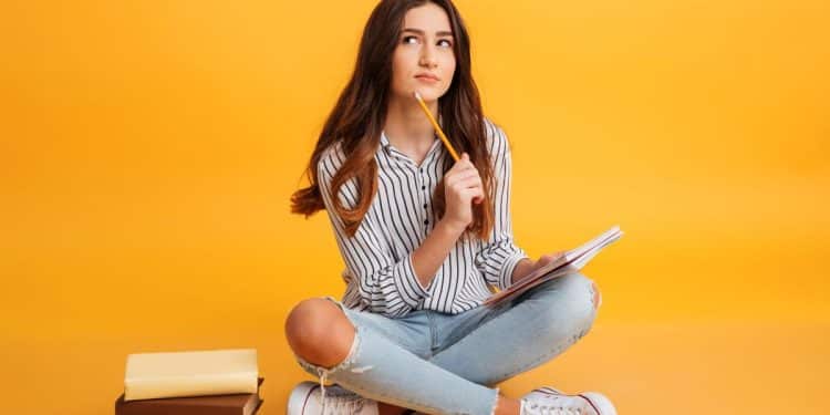 Jovem pensativa com caderno e lápis em fundo laranja.