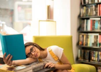 Estudante cansada apoiada sobre livros em biblioteca tentando se concentrar na leitura.