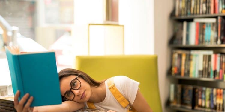 Estudante cansada apoiada sobre livros em biblioteca tentando se concentrar na leitura.