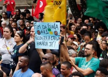 Manifestantes em praça pública segurando cartaz que diz "Fim da escala 6x1: reduza a carga horária já", representando o movimento pela mudança na jornada de trabalho no Brasil.