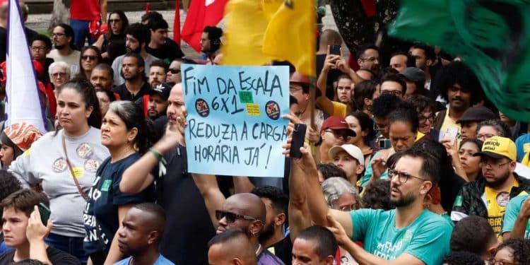 Manifestantes em praça pública segurando cartaz que diz "Fim da escala 6x1: reduza a carga horária já", representando o movimento pela mudança na jornada de trabalho no Brasil.