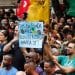 Manifestantes em praça pública segurando cartaz que diz "Fim da escala 6x1: reduza a carga horária já", representando o movimento pela mudança na jornada de trabalho no Brasil.
