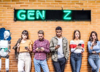 Grupo de jovens da Geração Z encostados em uma parede de tijolos laranjas, focados em seus celulares, ao lado de um robô humanoide. Acima deles, um letreiro neon escrito "GEN Z".