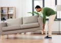 Homem levanta sofá bege em sala de estar moderna e iluminada.