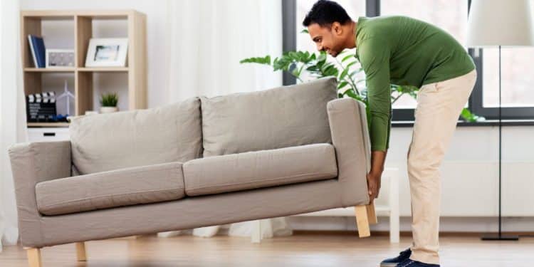 Homem levanta sofá bege em sala de estar moderna e iluminada.
