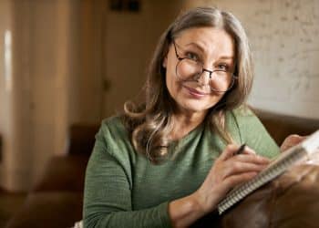 Mulher de cabelos grisalhos com óculos sorrindo enquanto segura um caderno em sofá marrom.