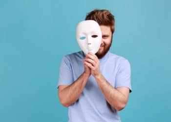 Homem segura máscara branca revelando olhar astuto por trás.