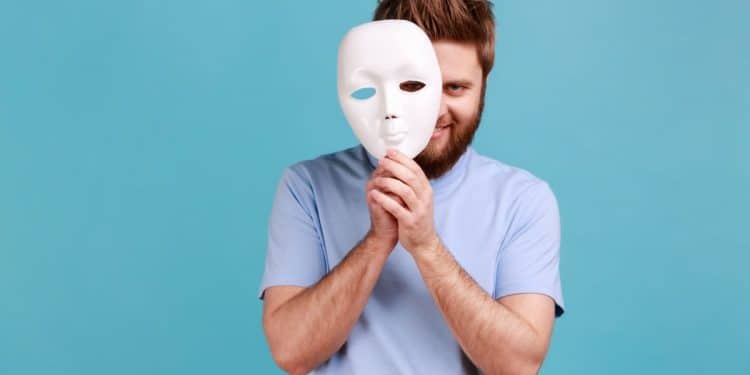 Homem segura máscara branca revelando olhar astuto por trás.