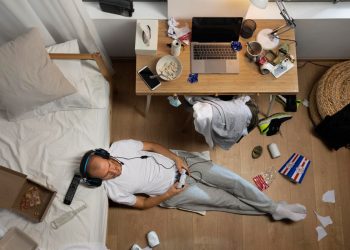 Homem jogando videogame no chão de um quarto bagunçado. Exemplo de comportamento habitual ligado ao sedentarismo e distrações.