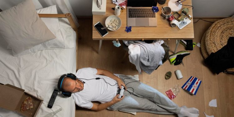 Homem jogando videogame no chão de um quarto bagunçado. Exemplo de comportamento habitual ligado ao sedentarismo e distrações.